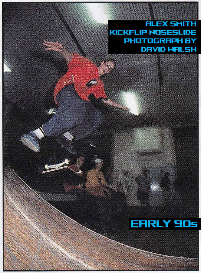 Alex mid-air doing a kickflip into a nose slide off a loading dock. Several Skaters in 90s Fashion watch in the background. 