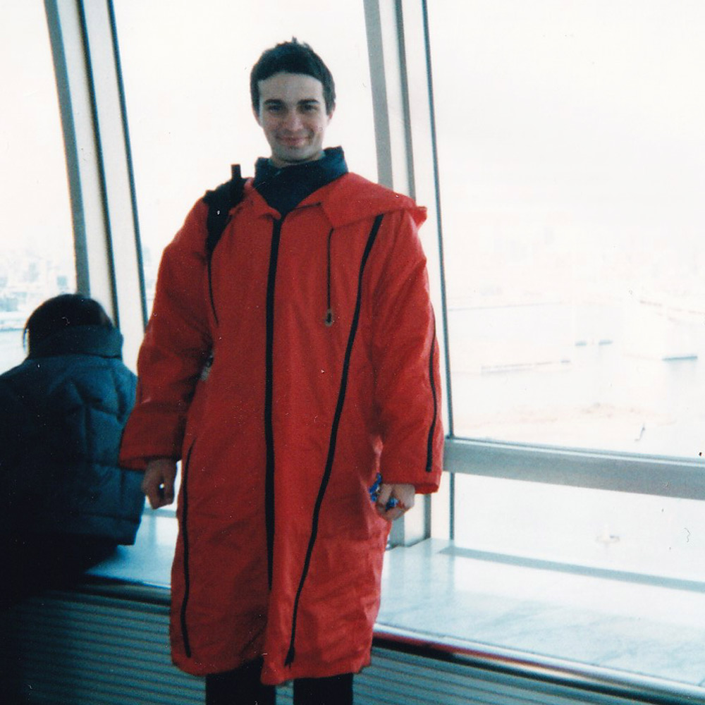 Image of Sean wearing a bright orange Final Home Jacket in front of a large window on a sunny, Winter day.
    Sean is smiling. The Final Home Coat is long - from shoulder to knee length with black vertical zippers featured vertically.
