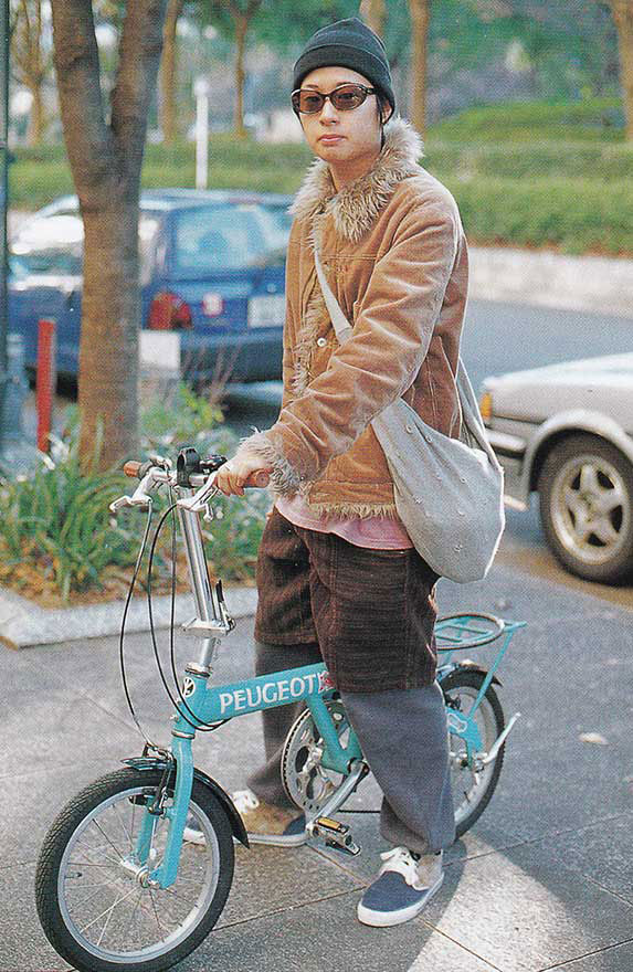 Photo of man with sunglasses and a beanie,
     standing above a Peugot Low Rider Bicycyle wearing Knot and Grand Canyon fashion brands.