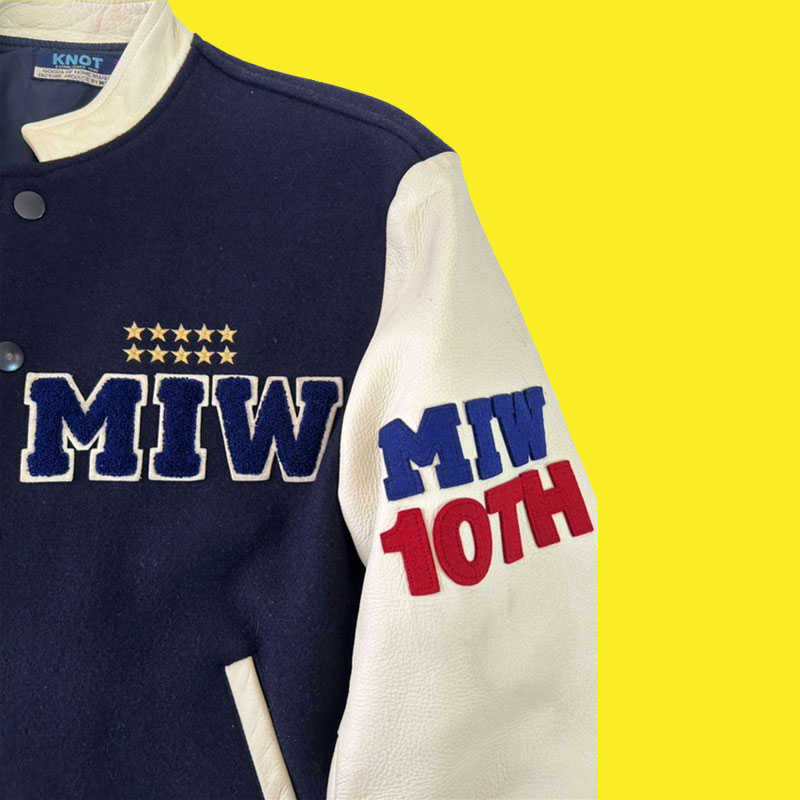 Close up image of vintage jacket by Knot - Made In World. The Jacket has white leather sleeves with
        a blue wool body piece - with applique reading 'MIW' and many embroidered stars above the logo.