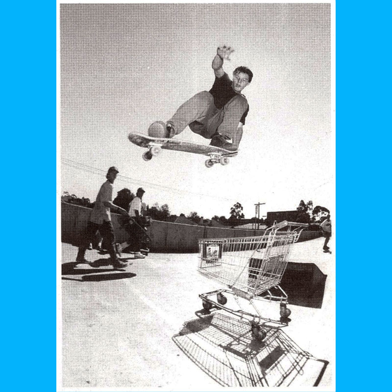 Jason Burrows launching off a jump ramp into the air - performing an Ollie Tail Grab high above (and over) 
    a supermarket trolley. Two skaters wearing baseball caps look on in amazement ih the background.