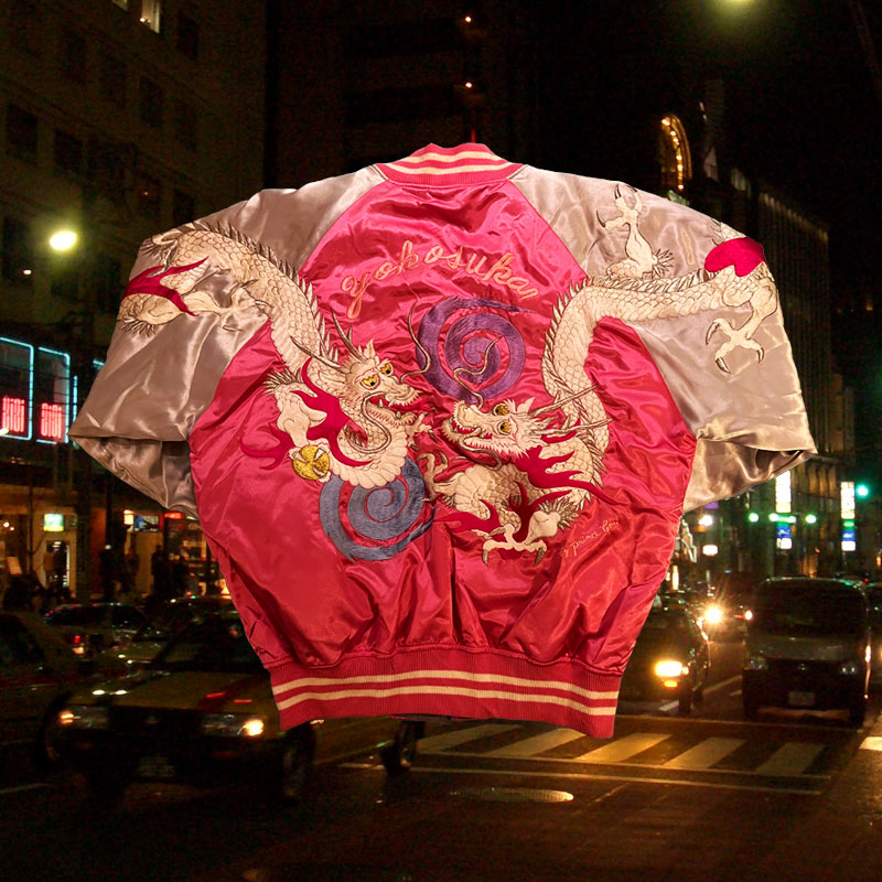 A very bright pink and silver Japanese Street Style Jacket with detailed embroidery of
    two dragons playing amongst circular motifs. The fabric is soft and silk like with the word 'Yokosuka' embroidered on the back.
    The background of the image is a busy Roppongi street with many cars.