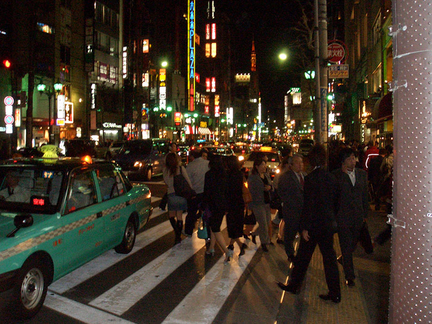 Photo of the main street in Roppongi on a busy weeknight. 
    A crowd of people are crossing the road with colorful neon lights in the background.