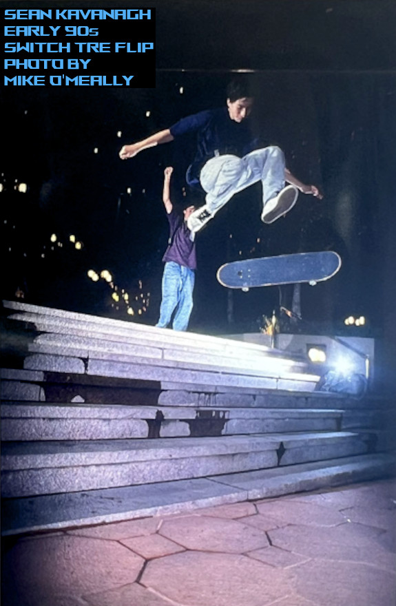 Photo of Sean doing a Switch Tre Flip down seven stairs late at night under the city lights. 
    A skater waves his hand high at the camera in the background.