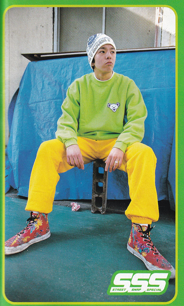 Photo of a man wearing Bright Green Sweater and Yellow Baggy Tracksuit Pants. 
    The sweater has an embroidered emblem of a bear character on the upper lapel area. He is a white and blue knitted beanie.