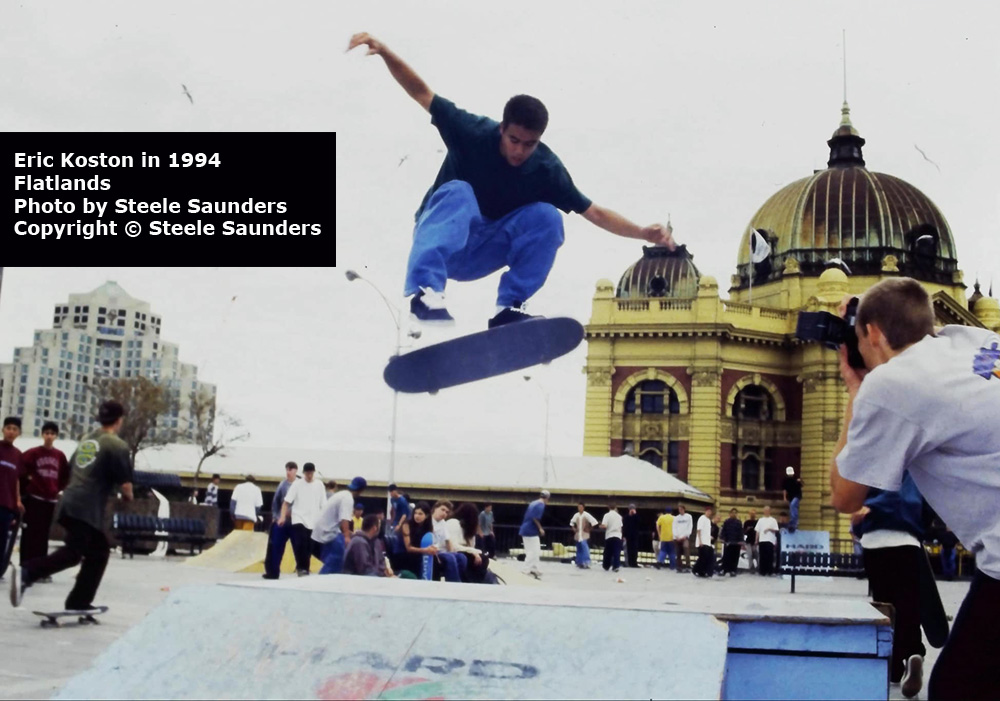 Eric Koston doing a Tre Flip over some ramps
        at the Flatlands Street Skateboarding Demonstration in 1994. There are skaters watching and hanging out
        in the background. Photo by Steele Saunders.